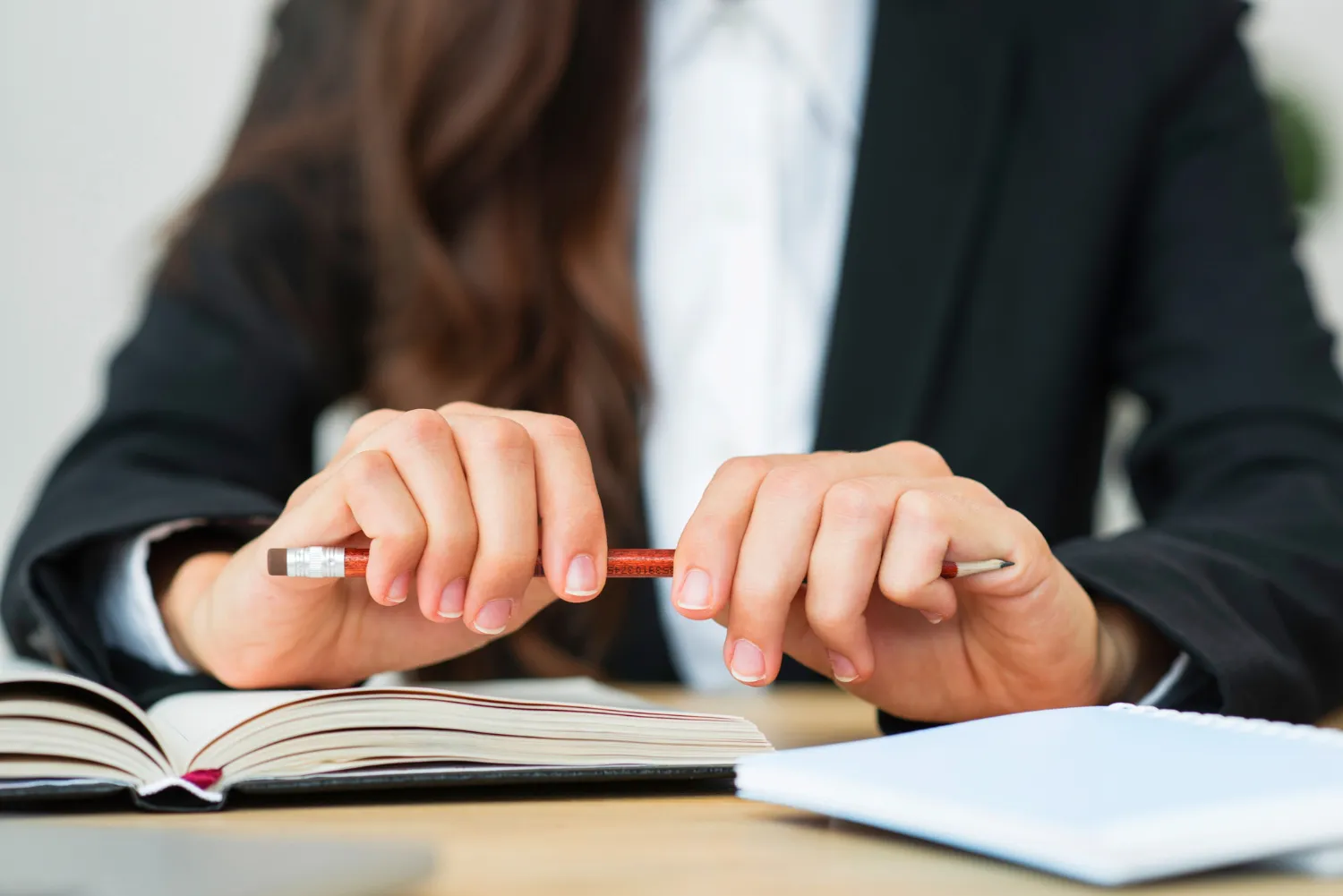 Mujer con traje de negocios sosteniendo un lápiz, sentada frente a un libro abierto y un cuaderno en una mesa de trabajo, en un ambiente profesional.